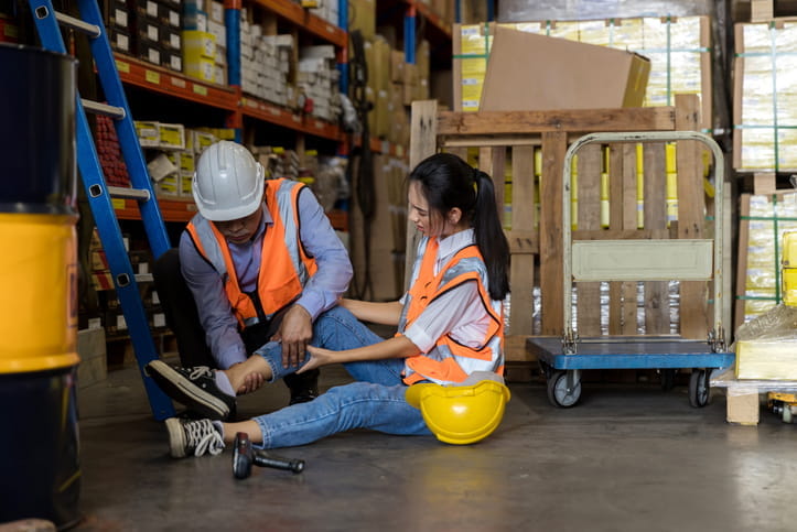 A male employee helps another employee who's injured on the floor of a warehouse, holding her leg in pain.