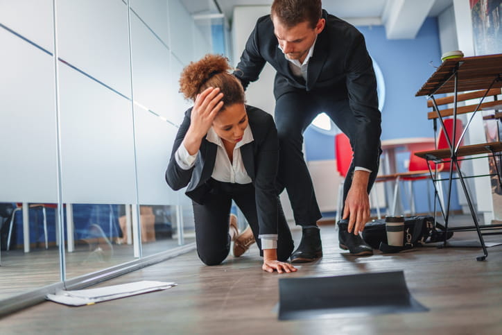 A male is helping his female coworker after she has slipped and fallen to the floor at work. Her belongings all scattered around the floor as she holds her head in pain.
